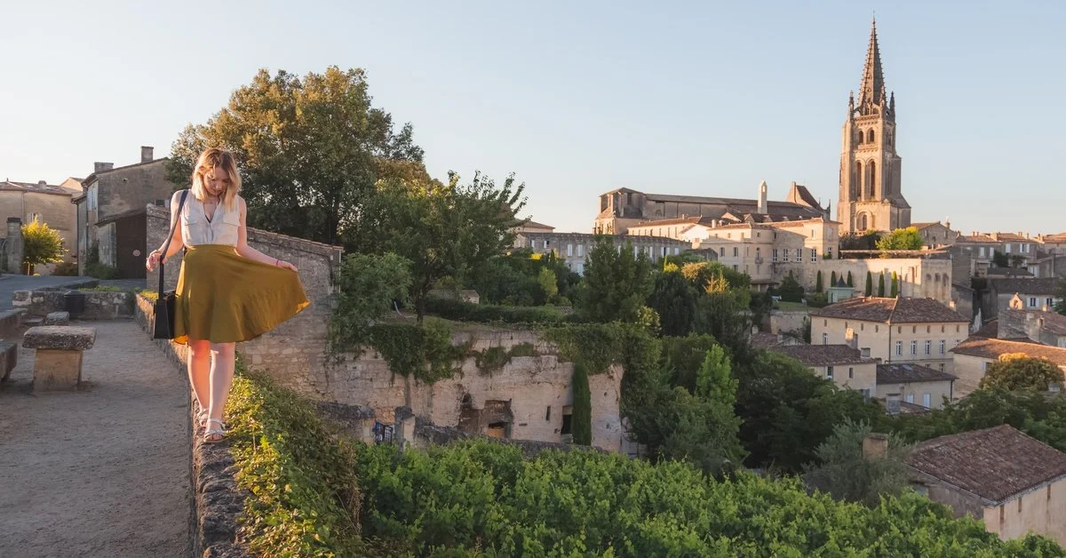 A woman walking along a stone wall overlooking a vineyard, old rooftops, and a tall church spire during golden hour.