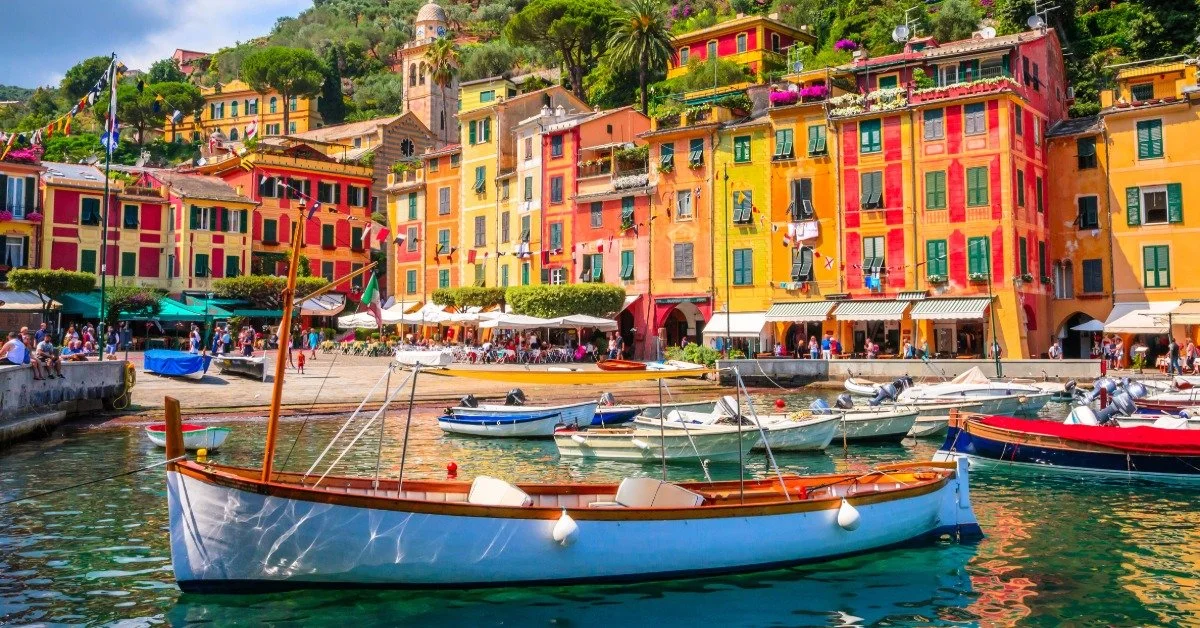 A row of tall, skinny, colorful homes line the coast of a waterfront town. Boats are in the water.