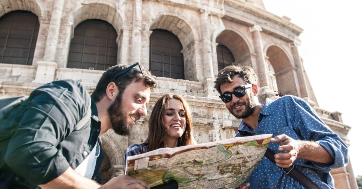 A smiling woman stands between two smiling men as they look at a map. The Roman Coliseum is behind them.