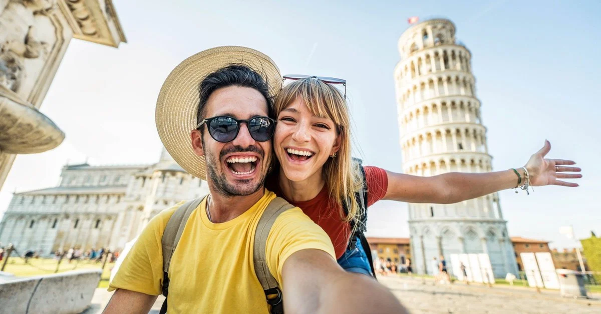A couple takes a selfie as they smile for the camera. They're in front of the Leaning Tower of Pisa in Italy.