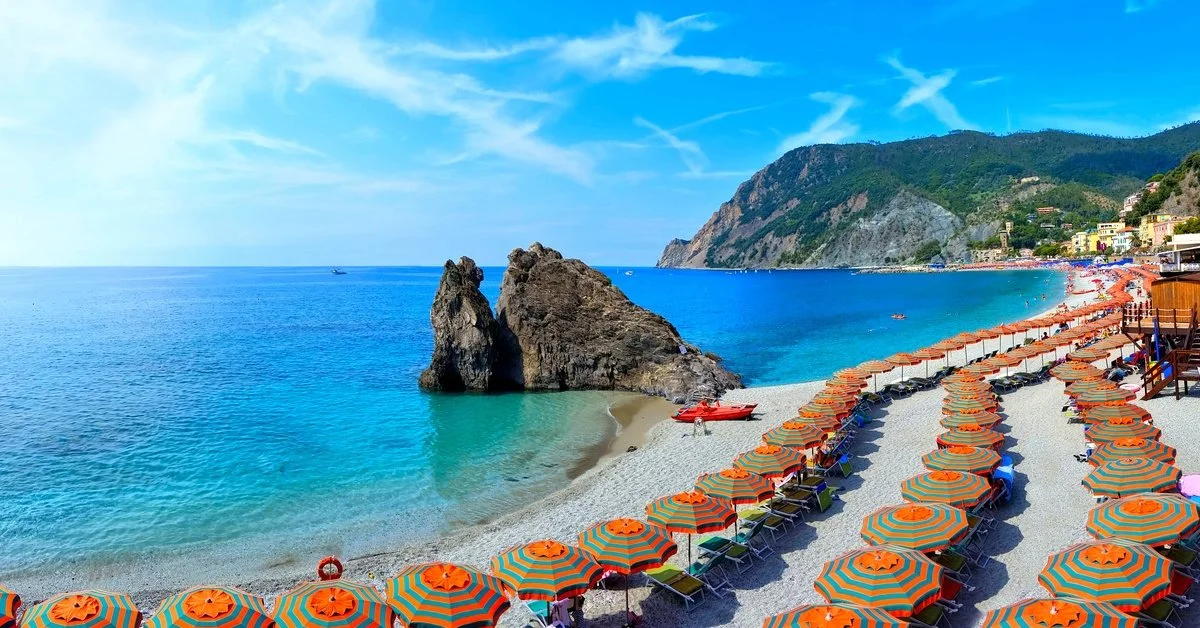 A panoramic view shows an Italian beach lines with orange and teal umbrellas on a sunny day. Mountains are in the background.