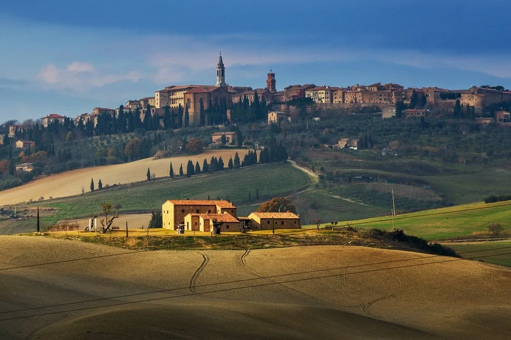 Scenic view of rolling hills with a small house and trees in the foreground, and a hilltop town with historic buildings and a church tower in the background.