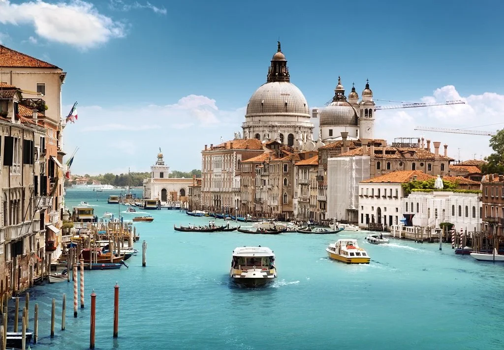 View of the Grand Canal in Venice, Italy, with boats, historic buildings, and the Basilica di Santa Maria della Salute in the background on a sunny day.