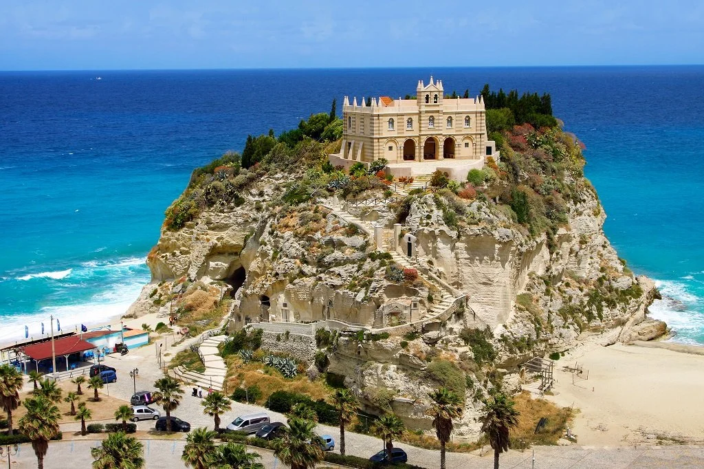 A castle on a rocky hill overlooking the ocean, with parking and palm trees at the base.