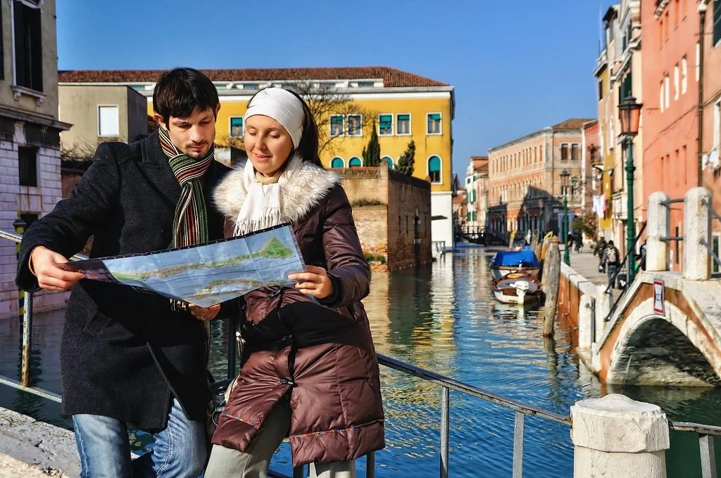 A young man and woman are standing on a canal side in Venice, Italy, looking at a map. The woman is dressed warmly in a coat and hat, while the man is in a dark coat and scarf. Colorful buildings line the canal behind them, with boats docked along the water.