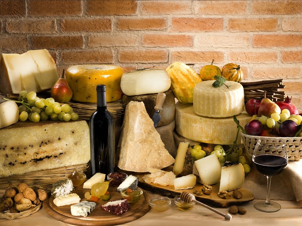 A variety of cheeses, grapes, a bottle of red wine, and assorted nuts and fruits arranged on a table against a brick wall.