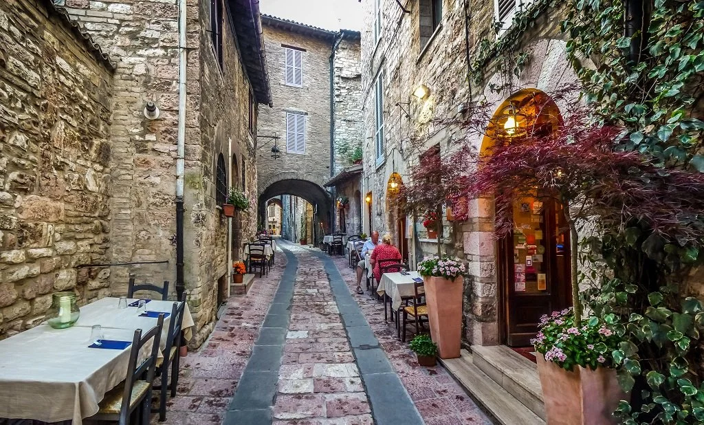 A narrow cobblestone street in a European village with outdoor restaurant seating, stone buildings, hanging lights, flowering plants, and an archway at the end.