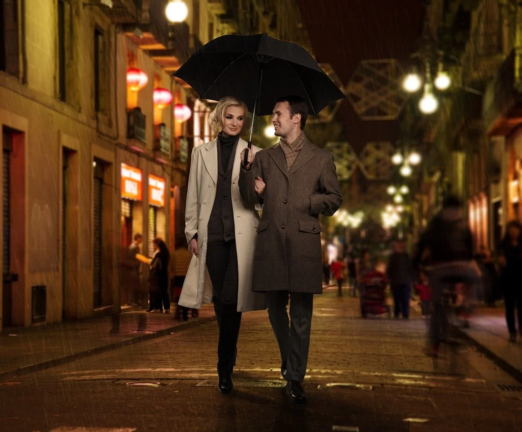 A couple walks arm-in-arm on a city street at night, sharing an umbrella and smiling at each other. The street is illuminated with warm lights and has blurred pedestrians.