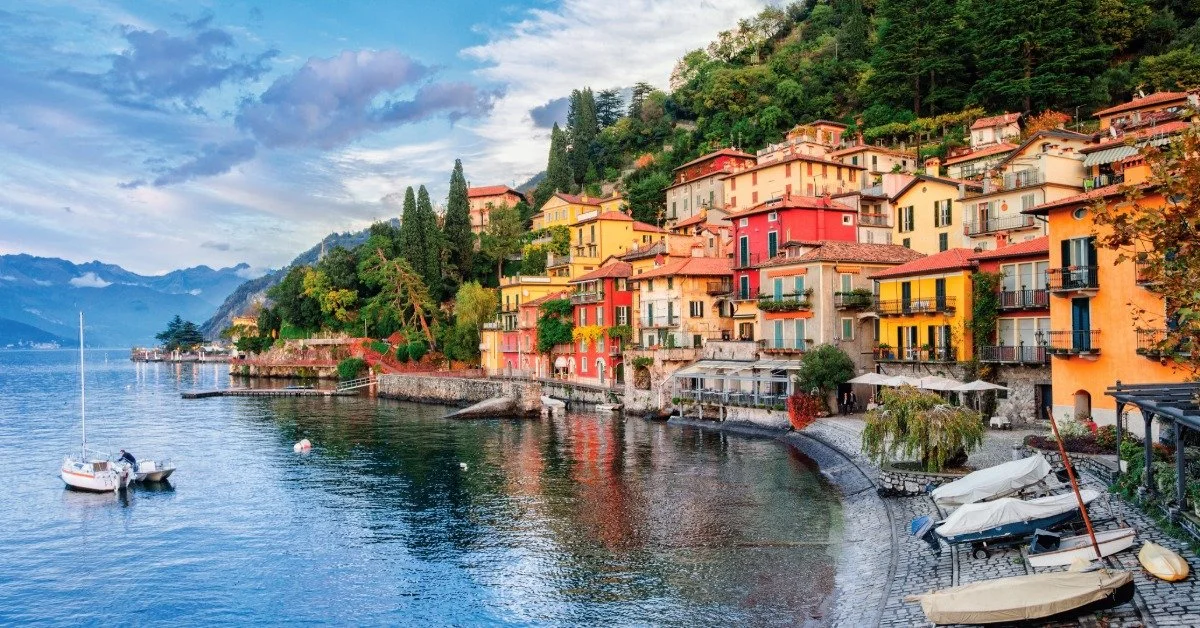 A village with tall yellow homes sits nestled into the hillside of mountains on one side and the shore of the lake on the other.