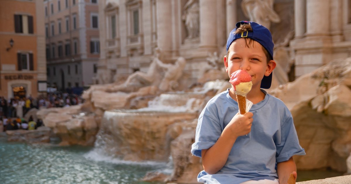 A young boy holds an ice cream cone while sitting on the edge of an old fountain. He's wearing a blue baseball cap backward.