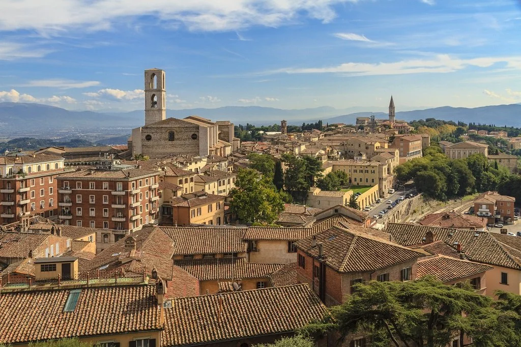 A panoramic view of an Italian city with historic buildings, church towers, and rolling hills in the background on a clear day.