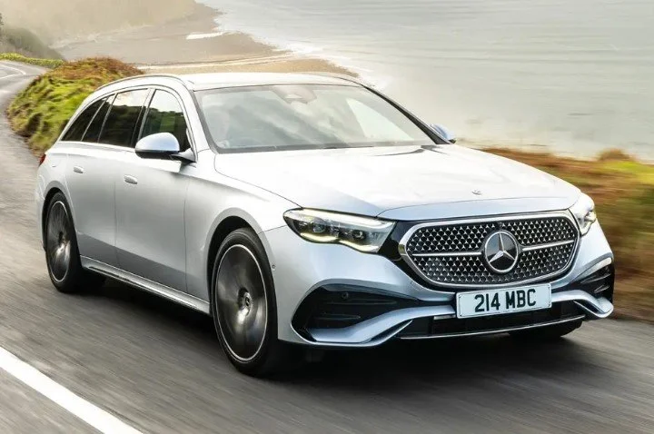 Silver Mercedes-Benz station wagon driving on a coastal road with ocean and grassy landscape in the background.