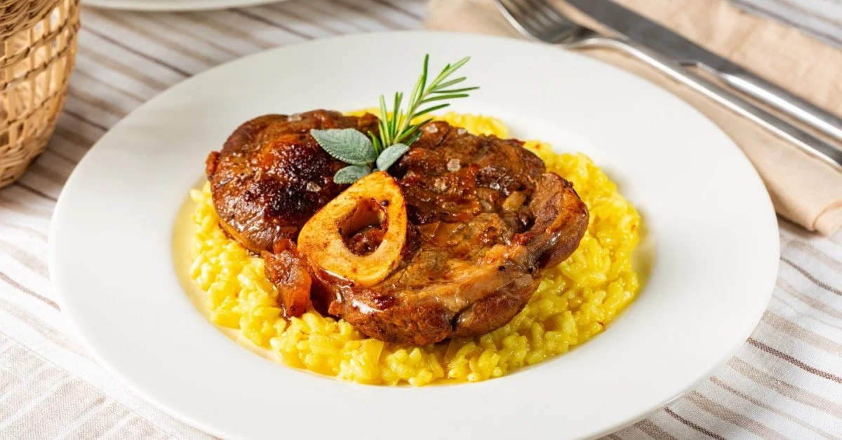 A white plate of ossobuco served over risotto alla Milanese and garnished with rosemary and sage.