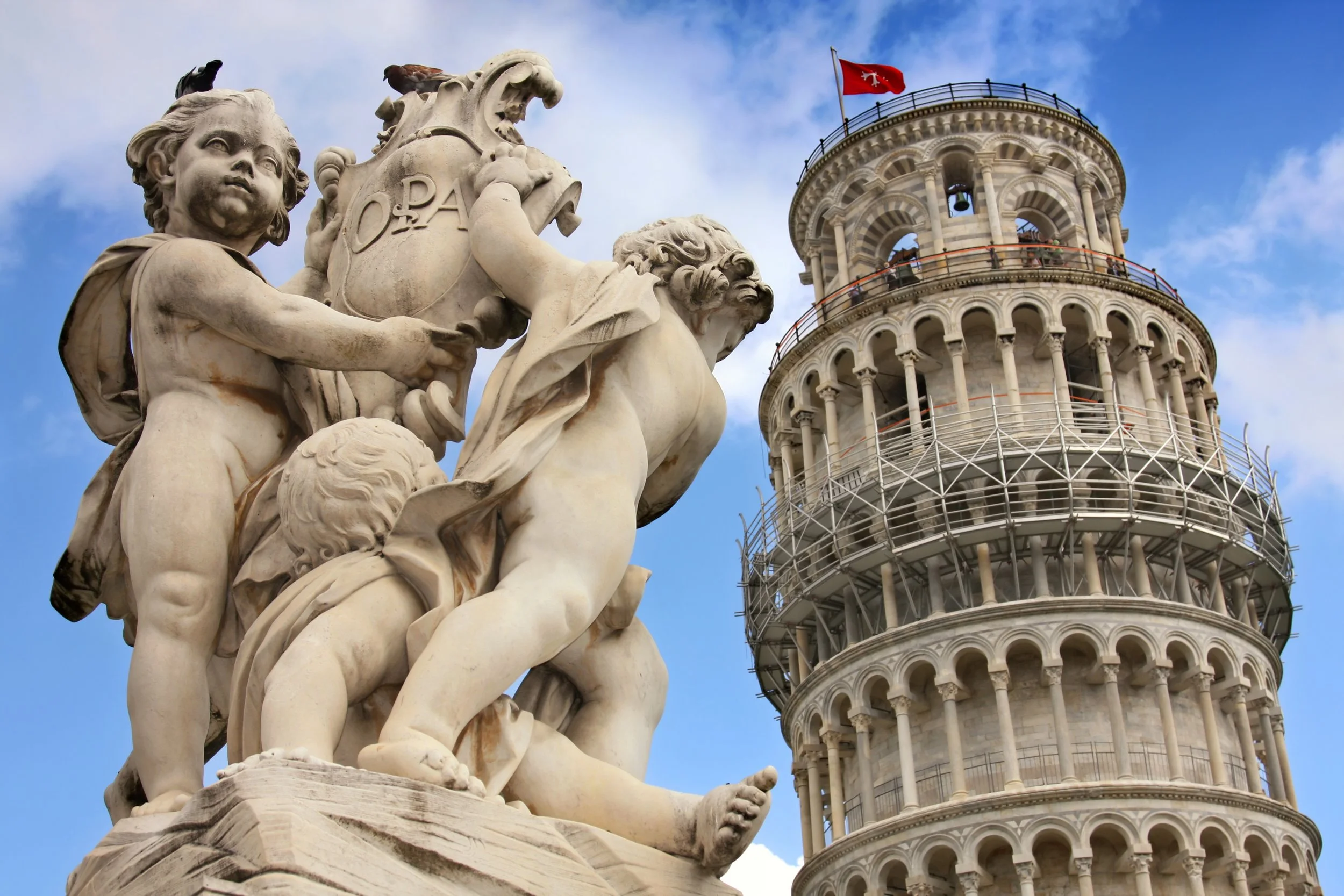 Close-up of marble sculptures depicting children and adults in front of the Leaning Tower of Pisa, with a blue sky and a red flag on top of the tower.