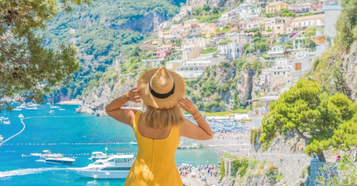 A woman wearing a yellow sundress and a tan, woven sunhat stands on a balcony looking at an Italian town.