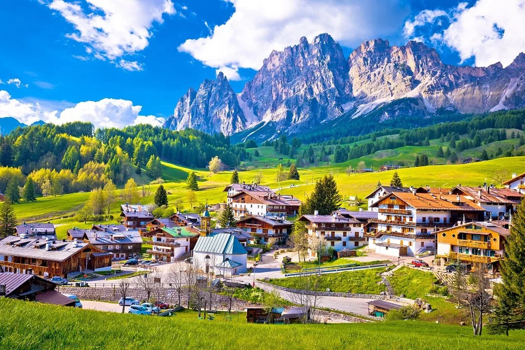 Cortina d'Ampezzo, an alpine village nestled in a lush green valley, with dramatic limestone peaks of the Dolomites in the background.