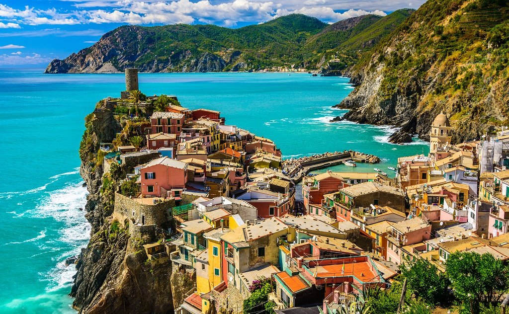 Colorful houses on a cliffside overlooking a turquoise sea with mountains in the background.