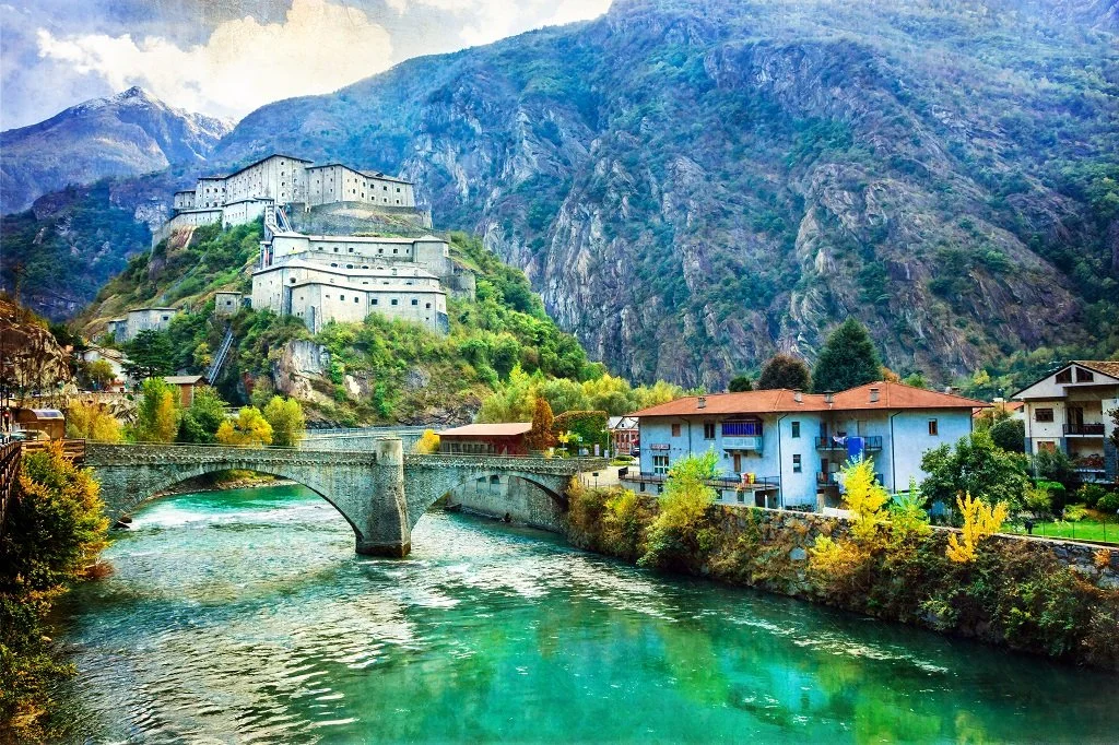 Scenic view of a historic castle on a hillside, with a bridge over a river and colorful buildings in a mountain village.