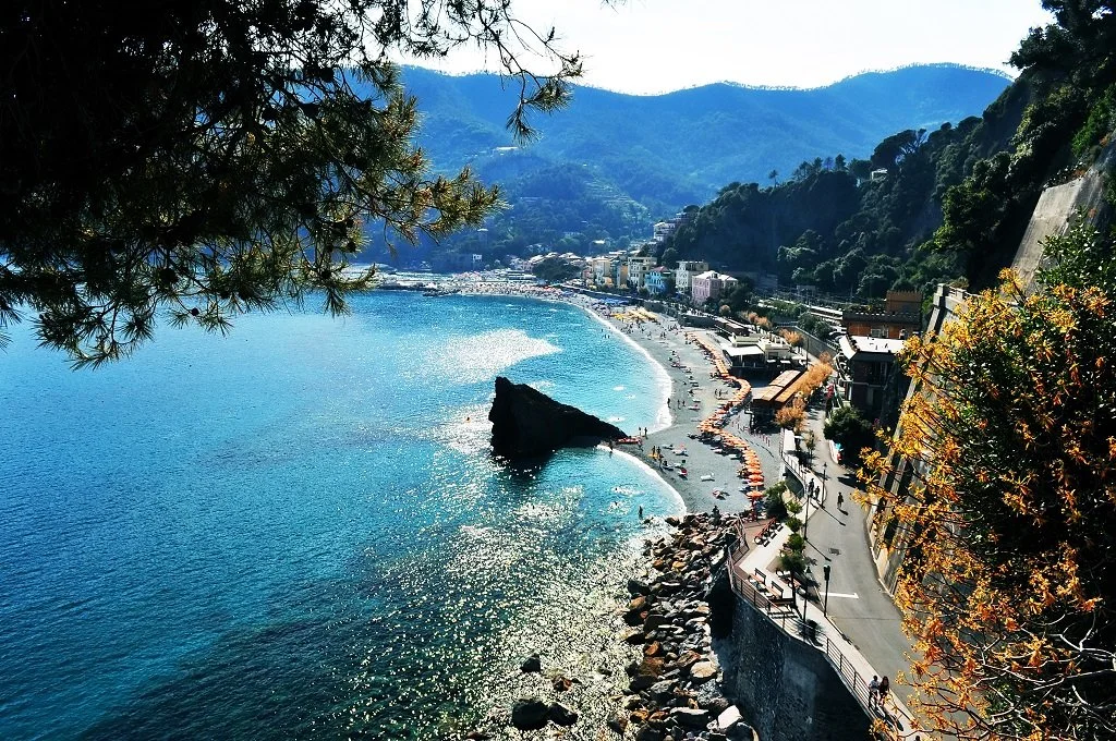 Scenic coastal view of a beach with a rocky promontory, surrounded by colorful buildings and a mountain backdrop, with trees in the foreground.