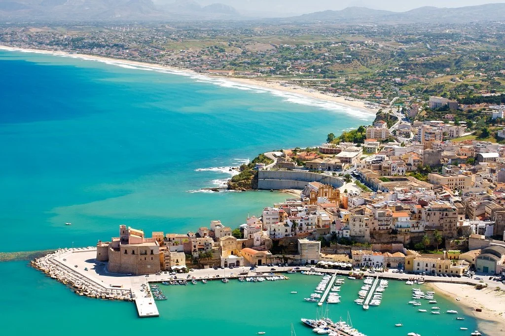 Aerial view of a coastal city with a marina, historic fortress, sandy beach, and turquoise waters, with a backdrop of city buildings and rolling hills.