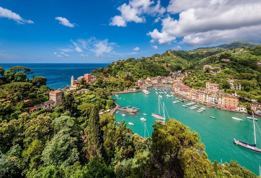 A scenic coastal town with colorful buildings along a marina filled with boats, surrounded by lush green hills and overlooking the ocean under partly cloudy skies.