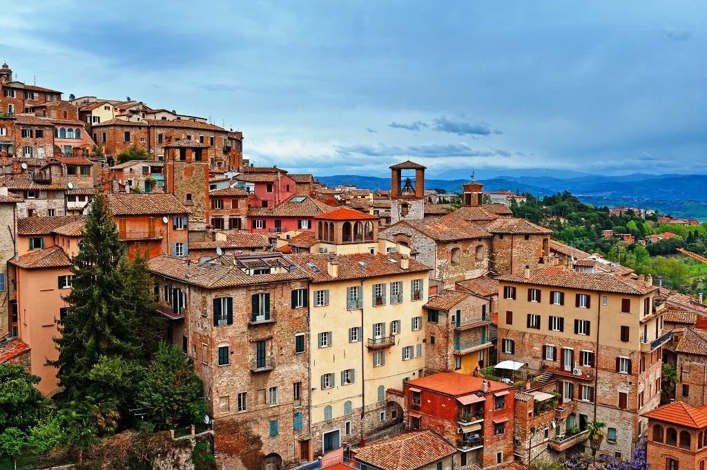 View of a hillside town with colorful, old-style buildings featuring terracotta roofs, set against a backdrop of rolling hills and cloudy sky.
