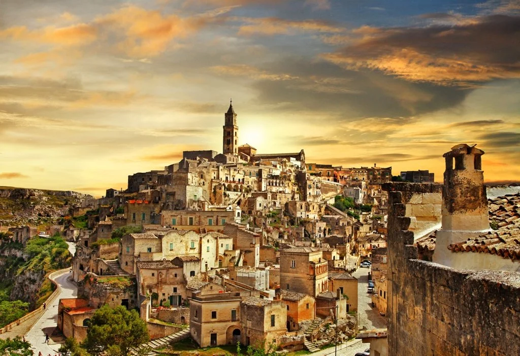 A hillside town with numerous old stone buildings and narrow streets, a tall church steeple in the center, during a sunset with golden and orange clouds in the sky.