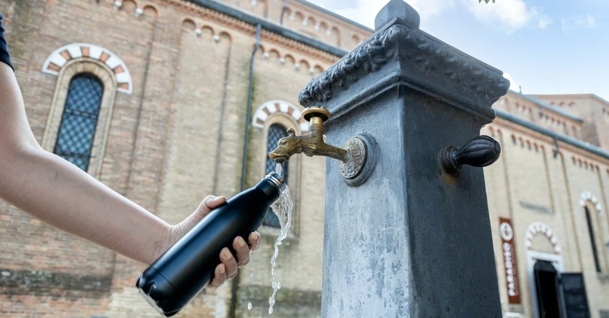 Someone filling up a black water bottle from a public Italian fountain in front of a brick church.