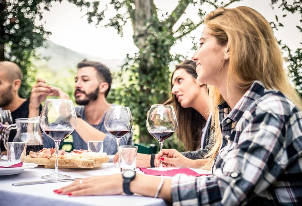 People sitting at an outdoor dinner table with wine glasses and a charcuterie board, enjoying a meal in a garden setting.