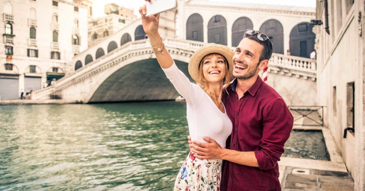 A young man and a young woman are standing next to a canal in Venice. They are taking a photo and smiling.