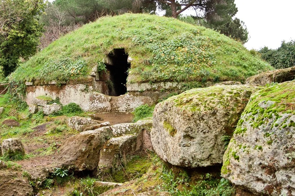 An ancient underground tomb built into a hillside, covered with grass and surrounded by large moss-covered rocks.