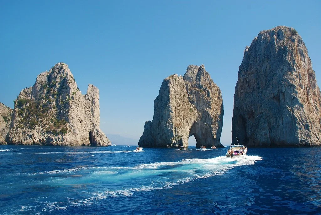 Boats sailing near large rock formations in the ocean under a clear blue sky.