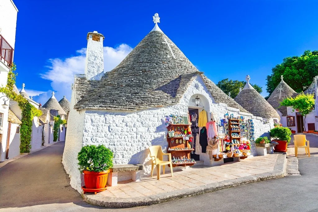 White stone buildings with conical roofs and small storefronts, possibly in a rural European village, with souvenirs on display outside.