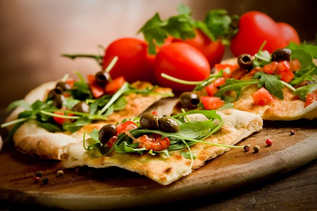Three flatbread pizzas topped with arugula, olives, chopped tomatoes, and slices of red tomatoes, placed on a wooden serving board with a bunch of red tomatoes and greenery in the background.