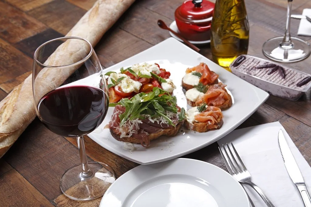 A glass of red wine, a basket of bread, a white plate with assorted appetizers including bruschetta with tomatoes and mozzarella, and other small spreads, on a wooden table with a fork and knife.