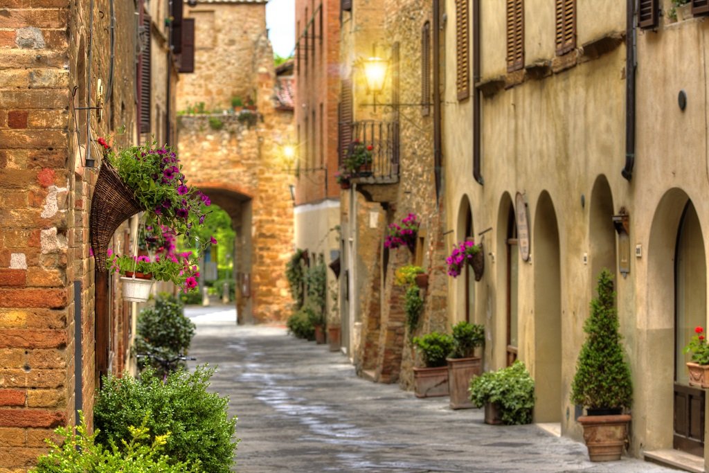 A narrow cobblestone street in a European village, with old brick and stucco buildings adorned with colorful flowers in window boxes and planters.