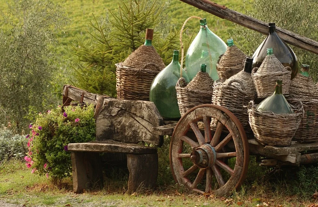 Old wooden cart filled with large glass bottles, some wrapped in wicker. The cart is situated outdoors near greenery and a wooden bench with flowering plants.