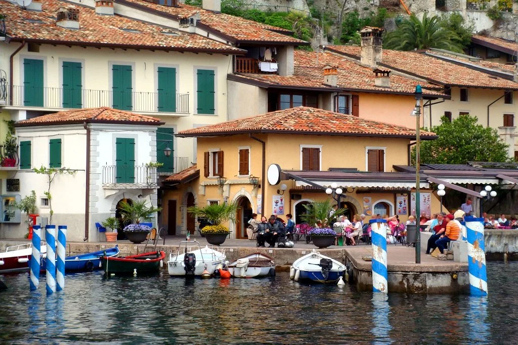 Colorful waterfront scene with boats docked at a pier, outdoor cafe with people sitting at tables, and buildings with green shutters and tiled roofs in the background.