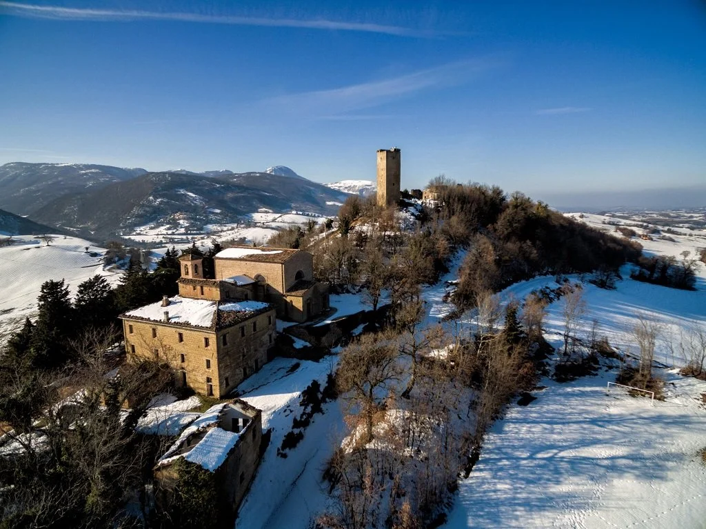 A snowy hilltop with old stone buildings and a tall tower, surrounded by trees and mountains in the distance under a clear blue sky.