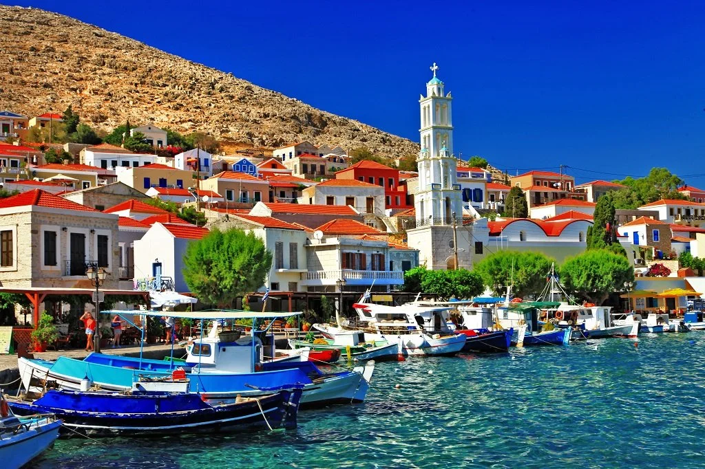 A scenic harbor with boats docked, colorful Mediterranean-style houses with red-tiled roofs behind them, a church with a tall white steeple, green trees, and a hillside under a clear blue sky.