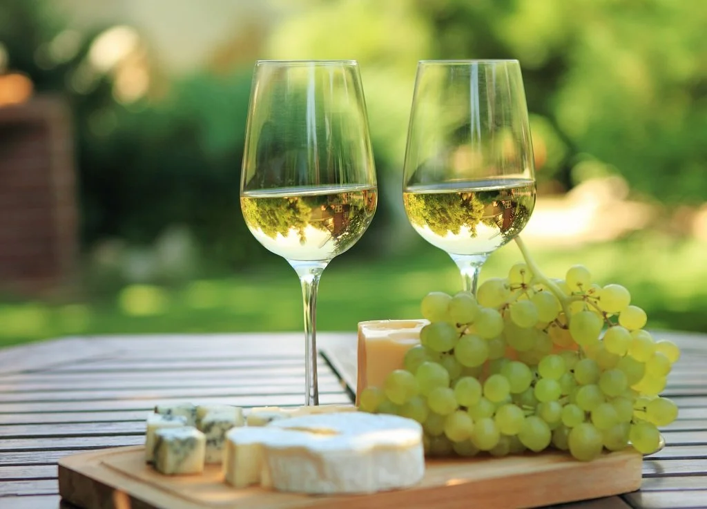 Two glasses of white wine, bunches of green grapes, assorted cheeses on a wooden board, and a candle on a table outdoors with a blurred green background.