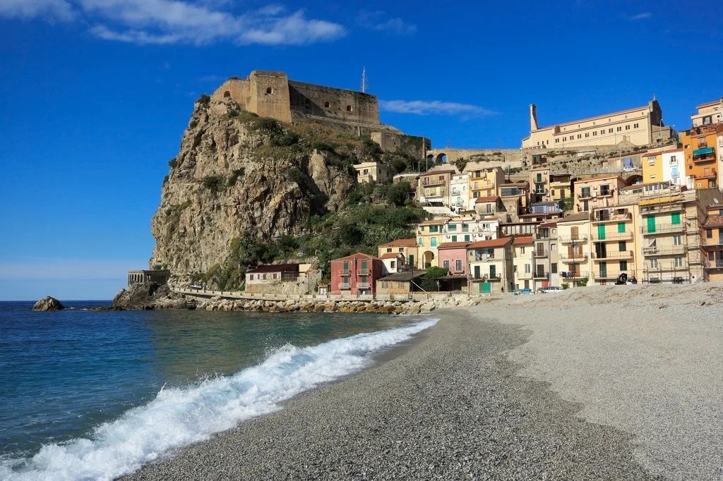 Coastal town with colorful buildings at the base of a cliff, a castle at the top, and a pebble beach with waves in the foreground