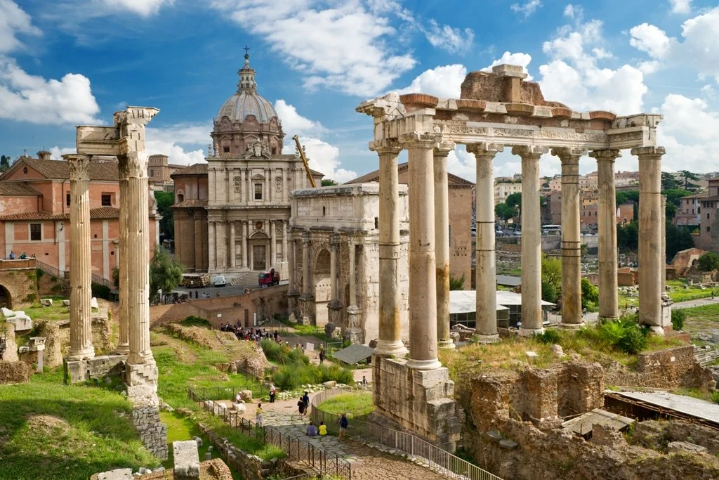 Ancient Roman ruins with columns and a church with a large dome in the background, under a partly cloudy sky.