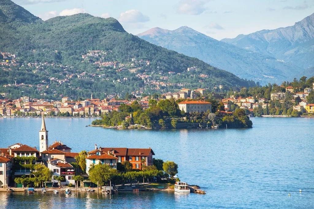 A scenic view of a lakeside town with colorful buildings, a church with a steeple, and lush green trees, surrounded by mountains in the background.
