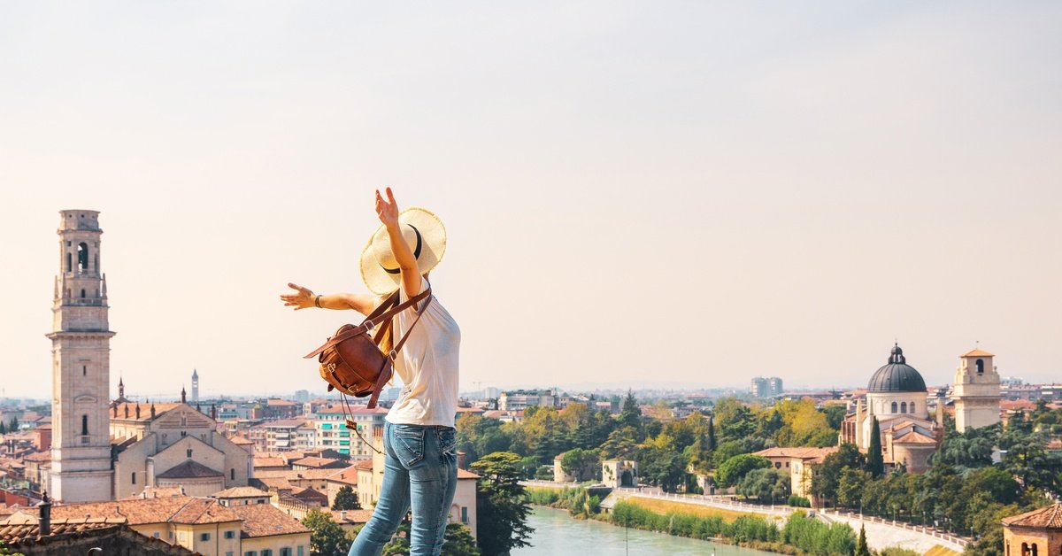 A woman in a sunhat joyfully flinging her arms back above a European river cityscape, framed by church towers and domes.