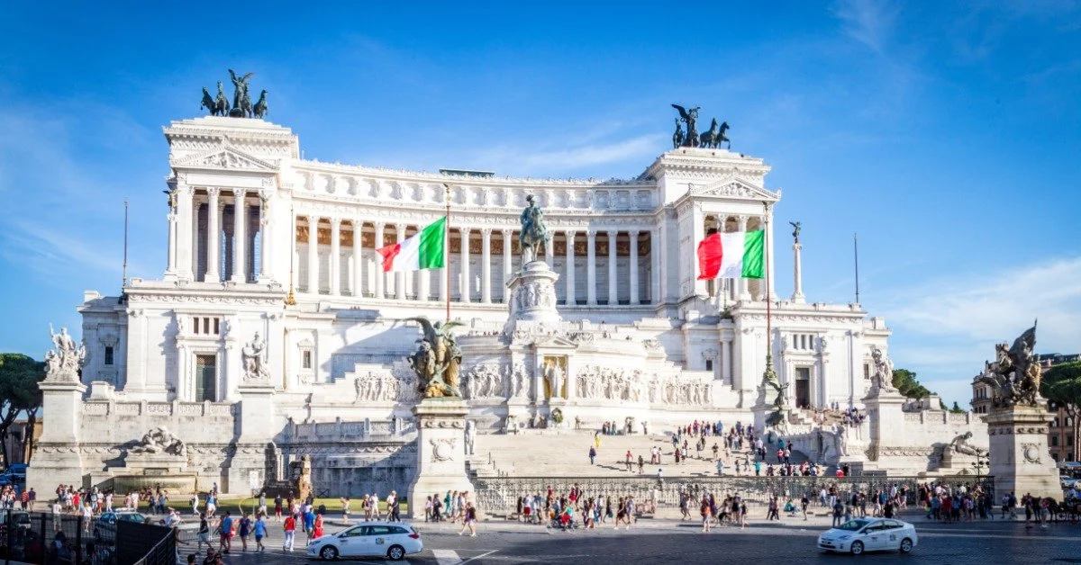 The Victor Emmanuel II Monument, with Italian flags and a large central monument, overlooks a busy square in bright daylight.