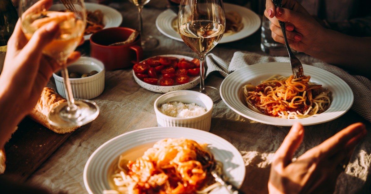 A close-up view shows people eating pasta from white plates and drinking wine from wine glasses.
