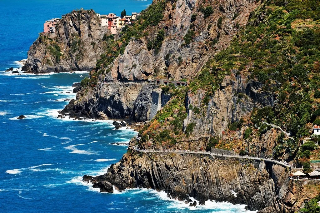 Steep coastal cliffs with a winding road and houses on top, overlooking the blue ocean with waves.