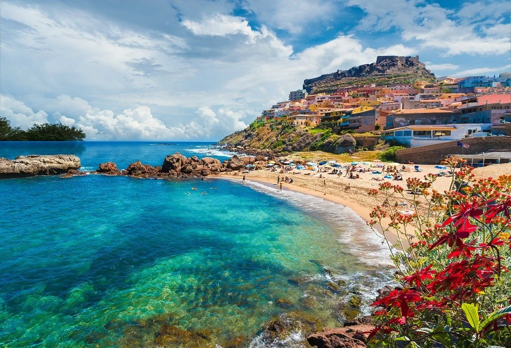 A coastal scene with clear turquoise water, a sandy beach filled with umbrellas and people, and colorful buildings on a hillside under a partly cloudy sky.
