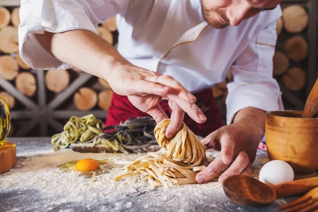 Chef preparing fresh pasta in a rustic kitchen with wooden logs in the background.